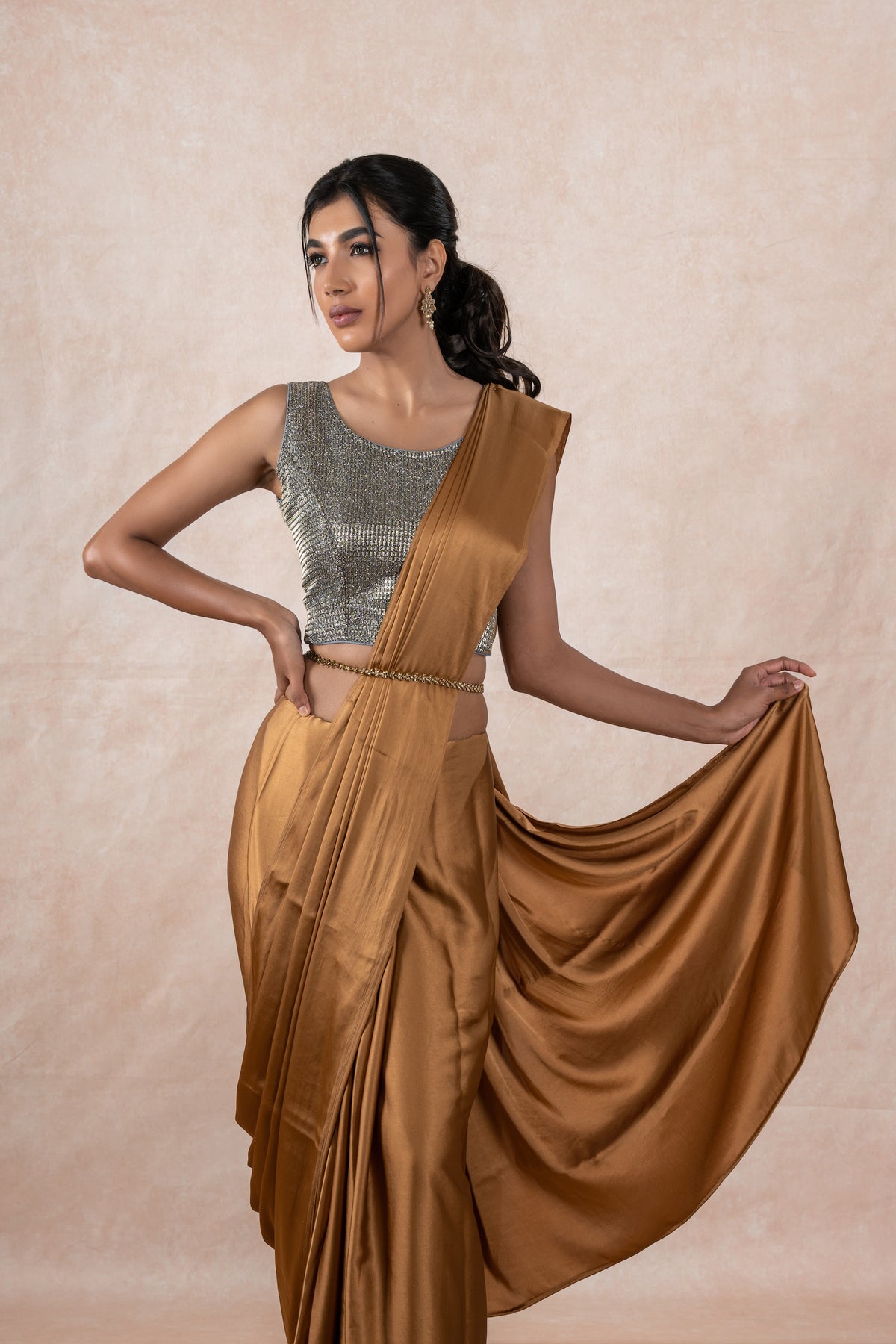 A model draped in a copper brown satin silk saree, grey shimmer blouse and gold buff saree shaper. The saree is pleated and  held with a waist chain.