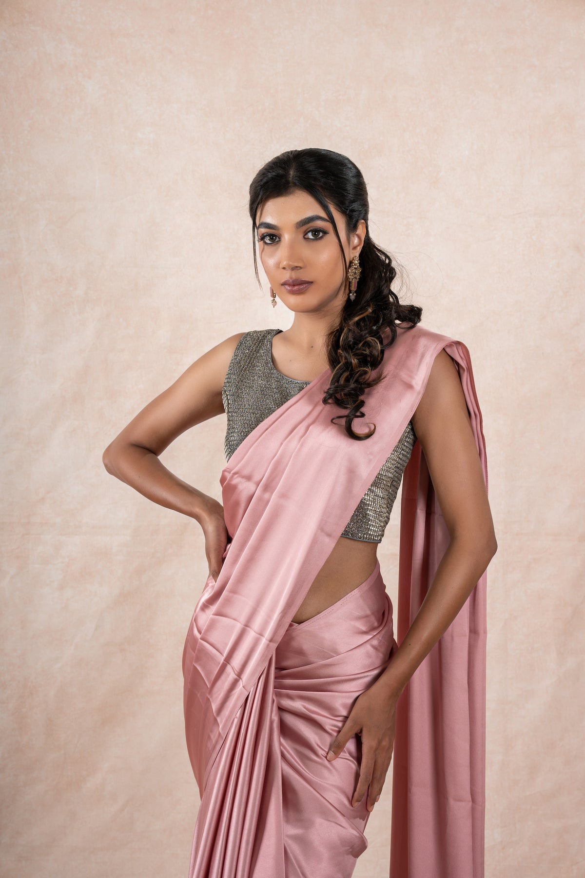 A model draped in a light pink satin silk saree, grey glitter blouse and light pink shaper. The saree is pleated beautifully around the shoulder.