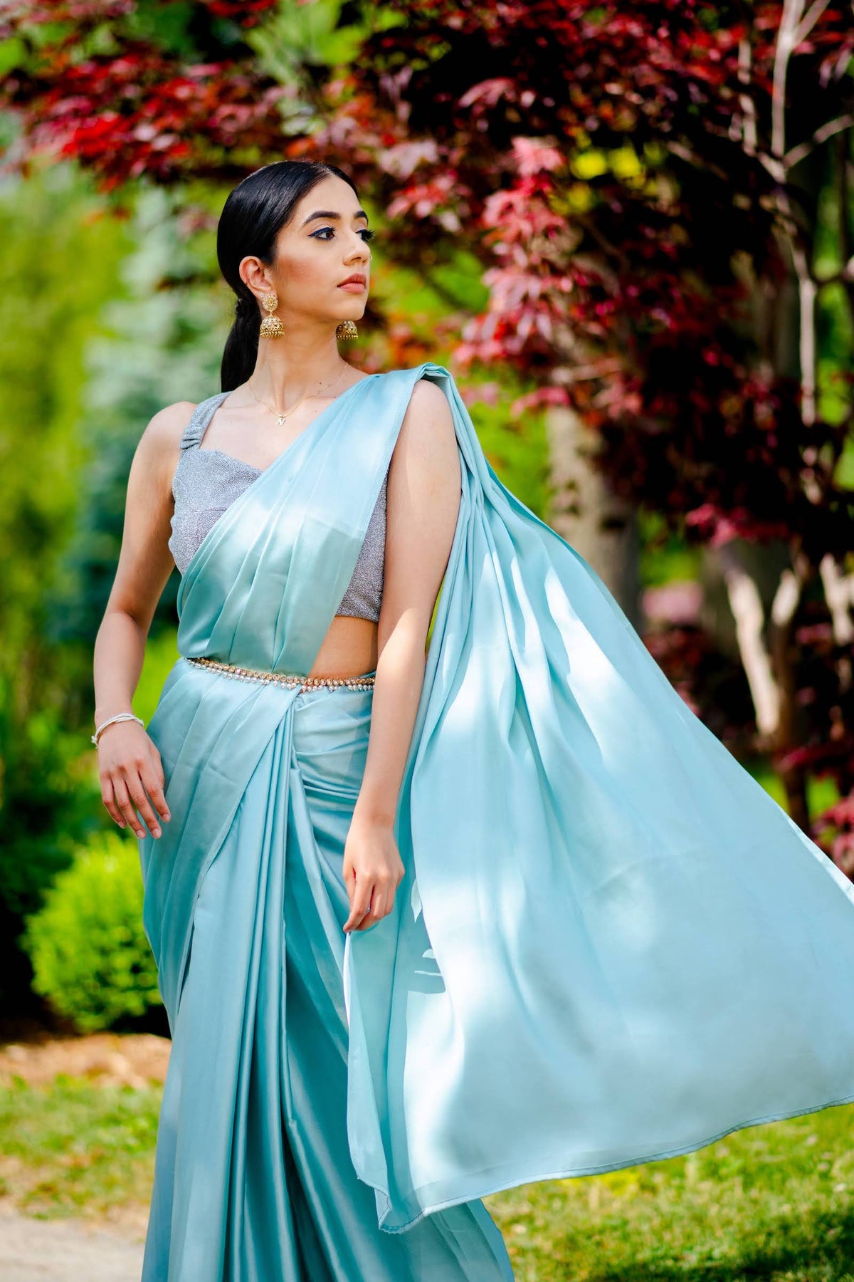 A model draped in an aqua blue satin silk saree, grey shimmer blouse and light blue saree shaper. The saree is pleated and  held with a waist chain.